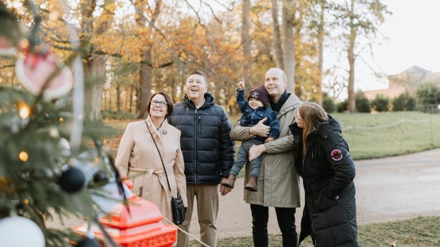 A group of four adults and a young child stand outdoors beside a decorated Christmas tree, looking at the festive display. The tree is adorned with baubles and lights, while autumn-coloured trees and parkland form the background. The child is being held by one of the adults and points towards the tree.